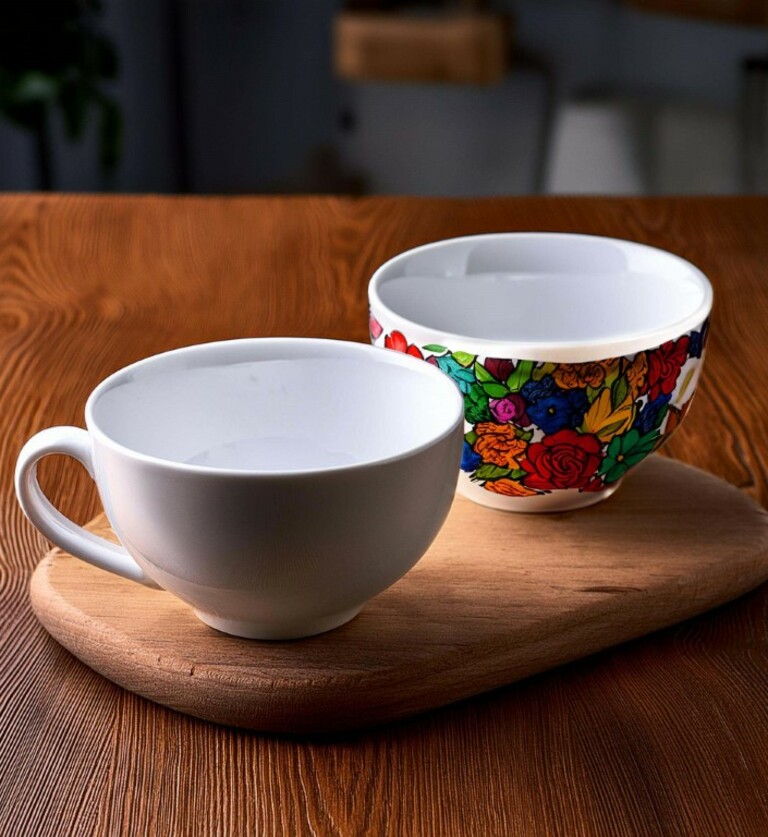 Zwei Tassen stehen auf einem Holzbrett auf einer braunen Holztischplatte. Die vordere Tasse ist unbemalt, die hinterer Tasse ist bunt bemalt.